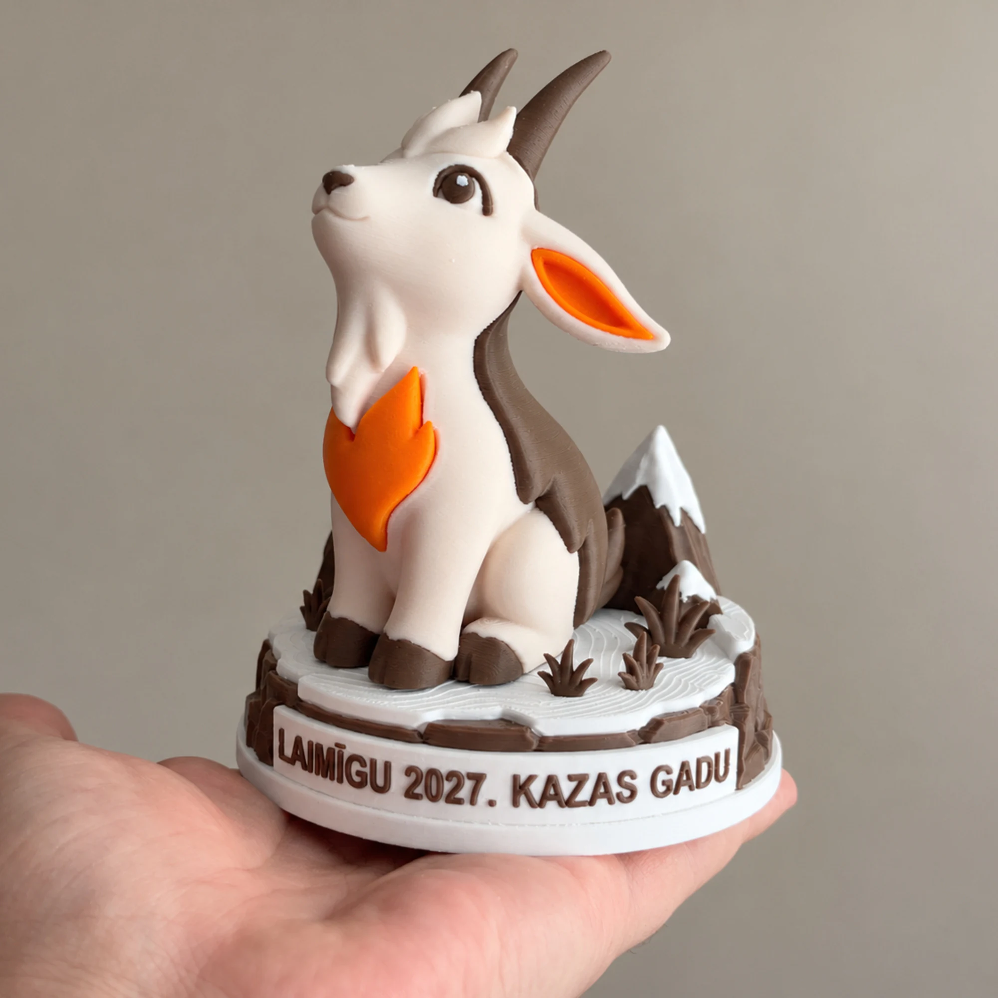 Goat figurine on its festive base in an adult palm, with the Latvian inscription LAIMĪGU 2027. KAZAS GADU.