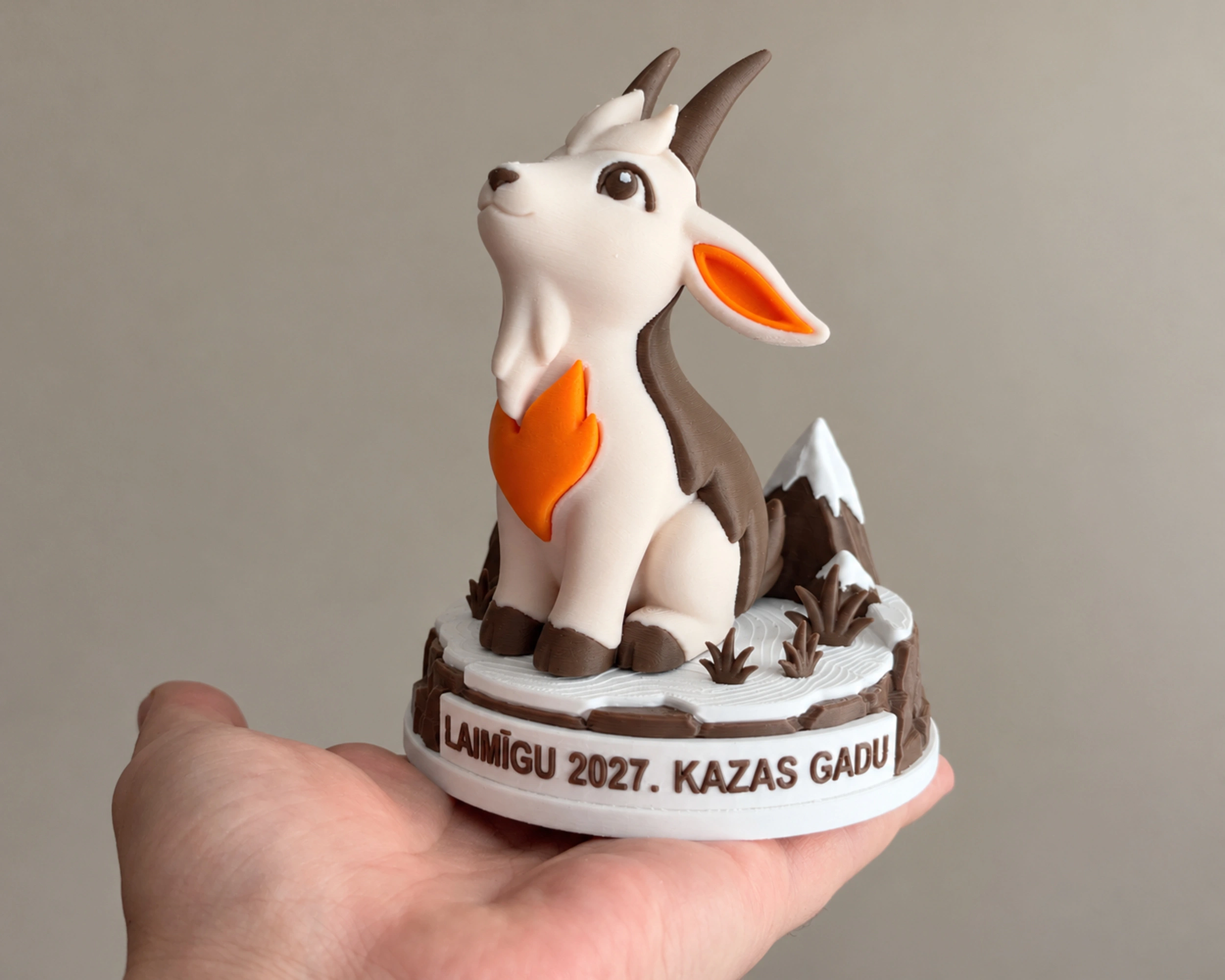 Goat figurine on its festive base in an adult palm, with the Latvian inscription LAIMĪGU 2027. KAZAS GADU.